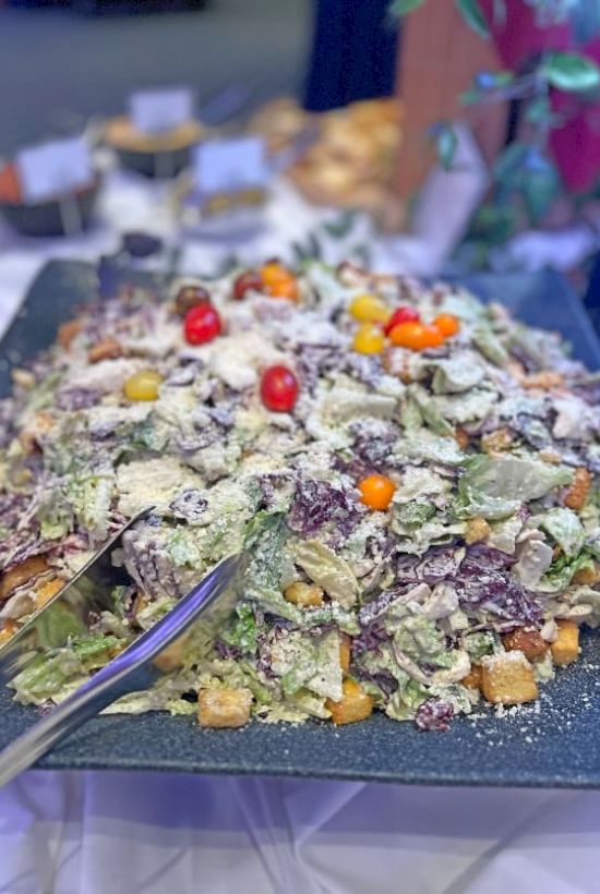A large, layered savory cake topped with greens, seeds, and small colorful vegetables, served on a black platter with a knife.