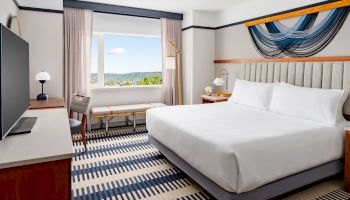 A bright hotel room with a king bed, bedside tables and lamps, a striped rug, a desk and chair by a window with a scenic view.