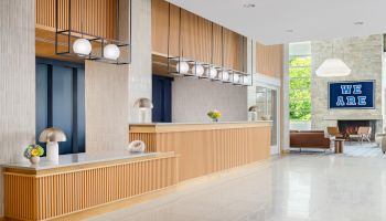 Modern hotel lobby with a long wooden reception desk, pendant lights, marble floor, and a seating area near a blue &ldquo;WE ARE HERE&rdquo; sign.