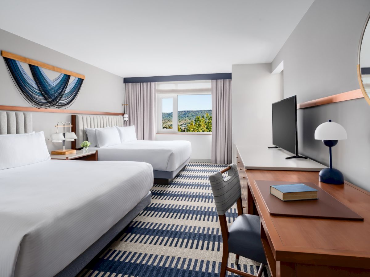 A hotel room with two large beds, a desk with a chair, a wall-mounted TV, a round mirror, and a window view of trees and sky, in a bright, modern space.