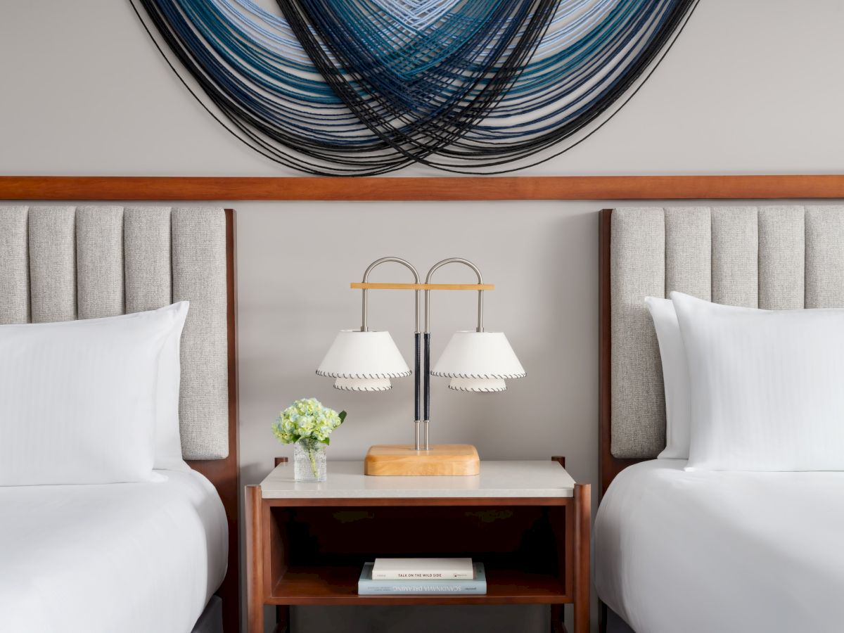Two hotel beds with white linens face a shared wooden nightstand between them, featuring a double lamp, a small plant, a book, and decorative wall art above.