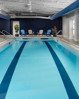 Indoor swimming pool with blue lanes, lounge chairs at far end, potted plants, large windows, bright lighting, and a calm atmosphere.