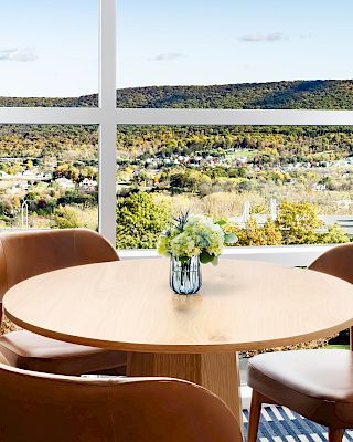 A cozy dining nook with a round wooden table, four brown chairs, a small vase of flowers, and a large window overlooking hills, curtains framing the view.