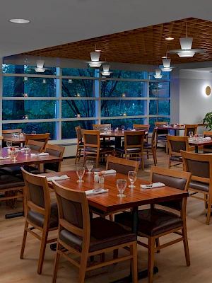 A modern dining room with wooden tables and chairs, large windows, plants, and soft lighting, ready for guests.