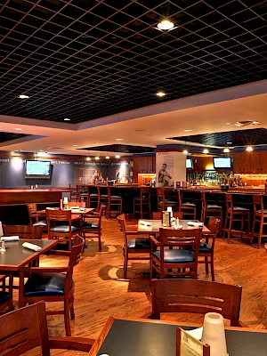 A spacious restaurant with wooden tables and chairs, dim lighting, and a bar in the back; empty tables set for service, casual dining vibe.