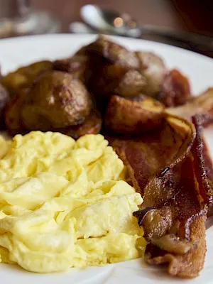 A plate of breakfast food: scrambled eggs, crispy bacon, a sausage, and sliced orange toast with a parsley garnish.