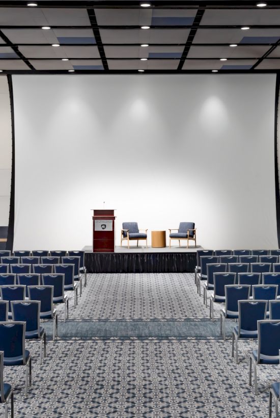 A large conference hall with rows of blue chairs facing a stage and a white projection screen, podium, and table at the front, empty and ready for a presentation.