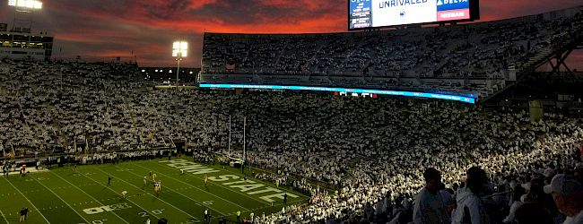 A football stadium at dusk with a fiery orange-pink sky, fans in the stands, and a bright jumbotron glowing above the field.