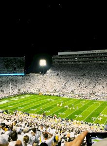 A nighttime stadium scene with bright field lights, empty stands, and a few people on the grassy pitch, under a dark sky.