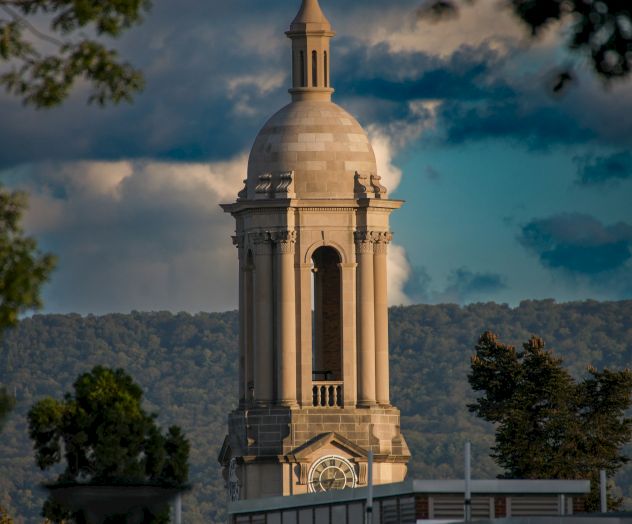 A tall, ornate monument with a spire rises above trees, under a dramatic cloudy sky. (140 characters)