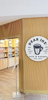 A caf&eacute; storefront with a wooden slat wall and a round Dear Joe sign featuring a coffee cup, inviting entry into the shop.
