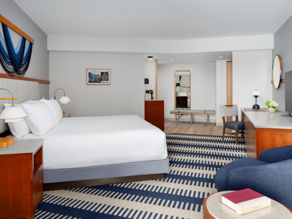 A modern hotel room with a large bed, wooden nightstands, a blue and white striped carpet, a desk, seating area, and a TV, all in neutral tones.