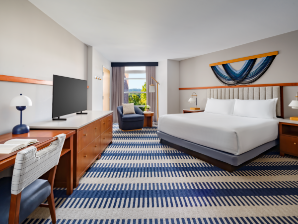 A bright hotel room with a king bed, desk, TV, and a seating area by the window, featuring blue striped carpet and nautical wall decor, tranquil setup.