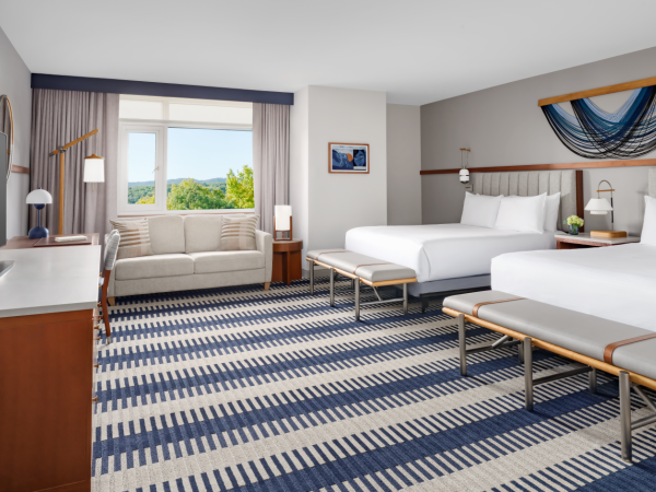 A hotel room with two beds, a seating area by a window, blue striped carpet, nightstands, lamps, and a bench at the foot of the beds, tropical vibe.