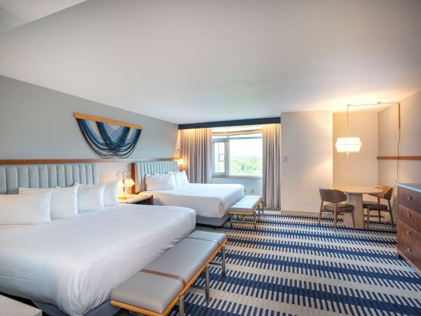 A bright hotel room with two queen beds, a striped blue rug, a wooden bench, seating area, desk, and large window with curtains.