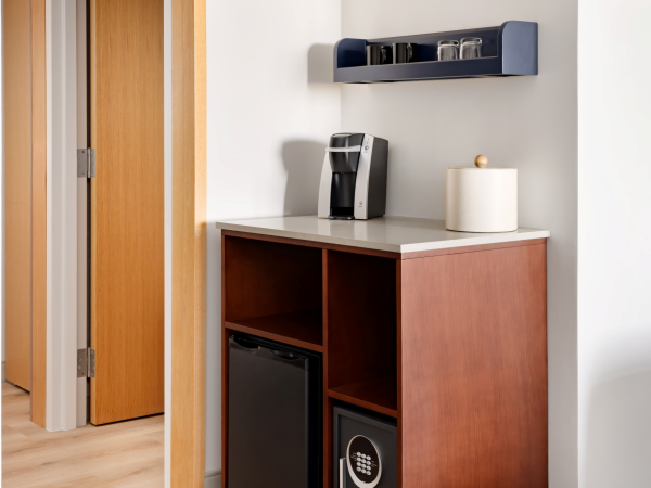 A compact coffee station with a countertop, a mini fridge and safe in a wooden cabinet, topped by a wall shelf and a coffee maker.
