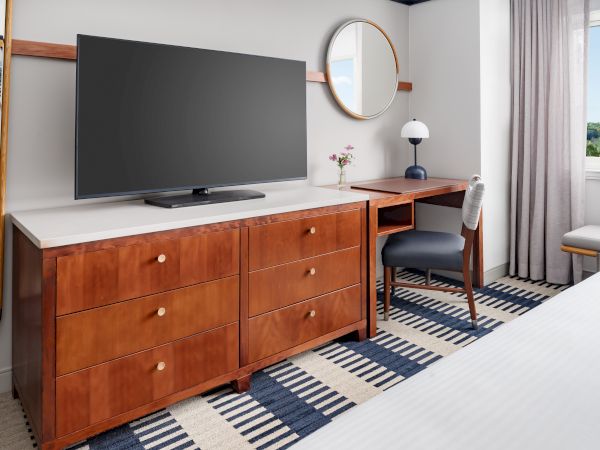 A modern hotel room with a wooden dresser, flat-screen TV, round wall mirror, desk and chair, bedside lamp, and a patterned rug by a window.