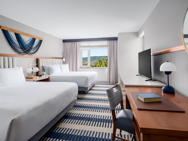 A hotel room with two beds, a desk, a flat-screen TV, a window with curtains, and a circular mirror; patterned carpet and blue accents.