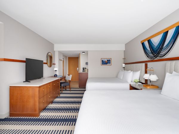 A bright hotel room with two large beds, a TV on a wooden dresser, a desk and chair, striped carpet, and wall art above the beds. Ends with a period.