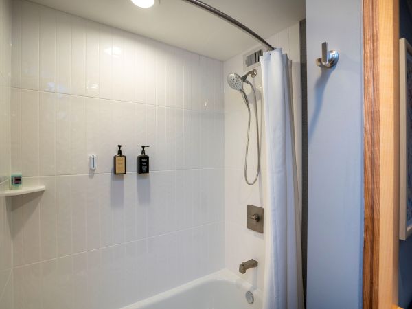 A white tiled bathroom shower with a glass door, handheld shower, wall-mounted soap/ shampoo bottles, a small shelf, and a curved ceiling light.