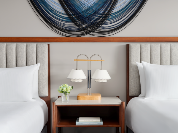 Two neatly made hotel beds with a shared nightstand, lamp, small plant, and a book underneath; modern art hangs above.