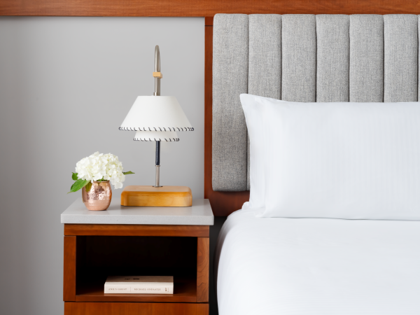 A neatly made bed with a gray upholstered headboard, a white duvet, and a bedside table featuring a lamp, a small plant, and a book.