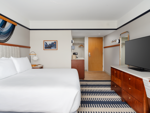 A bright hotel room with two white beds, a wooden desk and dresser, a flat-screen TV, striped carpet, wall art, and a doorway to a small kitchenette area.