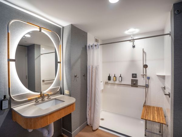 A modern bathroom with a backlit oval mirror over a wooden vanity, gray walls, and a glass shower with a wooden bench and shelves.