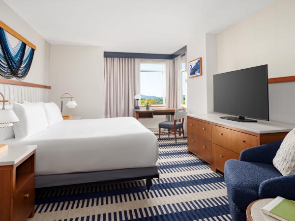A bright hotel room with a king bed, two lamps, a blue striped carpet, a desk near a window, a flat-screen TV on a wooden dresser, and a blue armchair with a cushion.