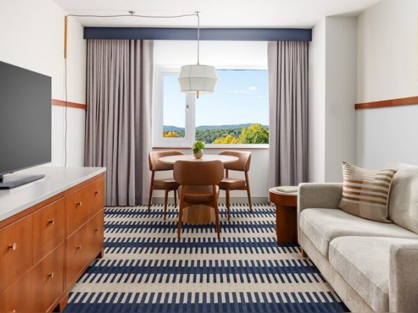 A hotel suite with a TV on a wooden dresser, a striped carpet, a beige sofa, a coffee table, and a dining area by a large window with curtains.