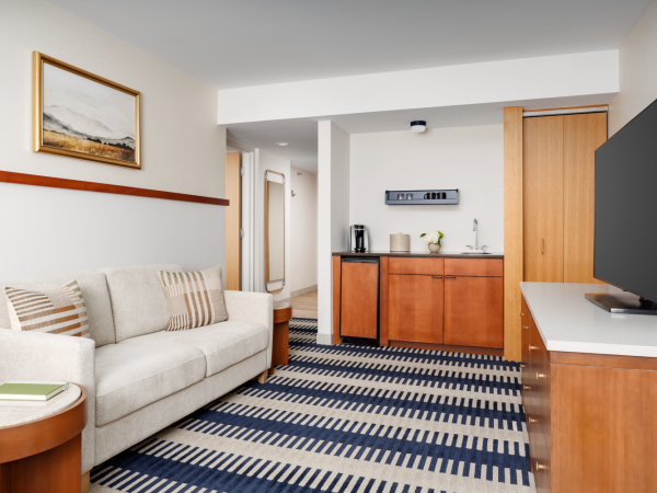 Cozy hotel suite with a beige sofa, striped rug, wooden furnishings, a small kitchenette, flat-screen TV, and a framed wall art piece.