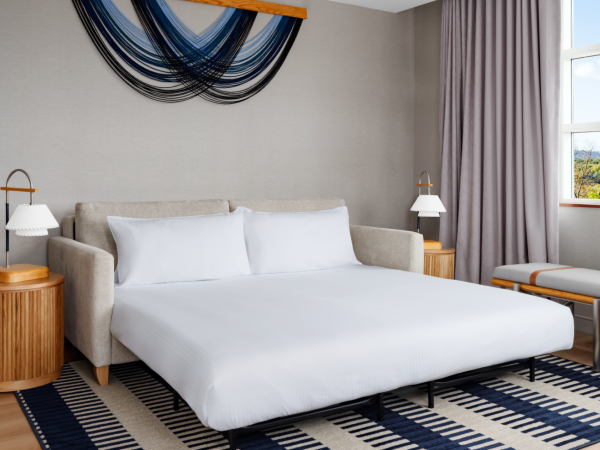 A modern hotel room with a white bed, beige sofa, two nightstands, lamps, patterned rug, and a window with purple curtains.