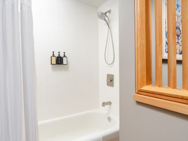 A bathroom scene with a white tub and shower, a white curtain, wall-mounted shampoo bottles, a handheld showerhead, and a wooden half-wall divider.