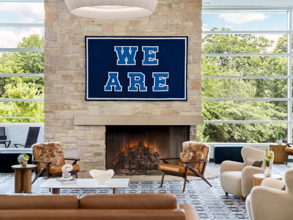 A cozy modern lounge with a stone fireplace, large windows overlooking greenery, and a bold &ldquo;WE ARE&rdquo; sign above the fire.