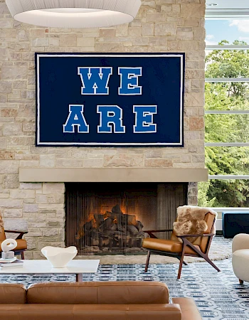 A modern lounge with a cozy fireplace, stone wall, large windows, and stylish chairs; a bold blue &ldquo;WE ARE&rdquo; sign above the fireplace, inviting atmosphere.