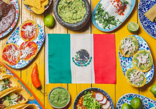 A colorful Mexican feast scene around a central Mexican flag, featuring tacos, tostadas, enchiladas, guacamole, salsas, quesadillas, lime accents, and vibrant plates.