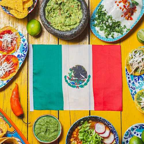 A colorful Mexican feast scene around a central Mexican flag, featuring tacos, tostadas, enchiladas, guacamole, salsas, quesadillas, lime accents, and vibrant plates.