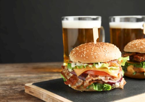 Two burgers with lettuce, tomato, cheese and bacon on a wooden board, plus two mugs of beer in the background.