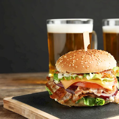 Two burgers with lettuce, tomato, cheese and bacon on a wooden board, plus two mugs of beer in the background.