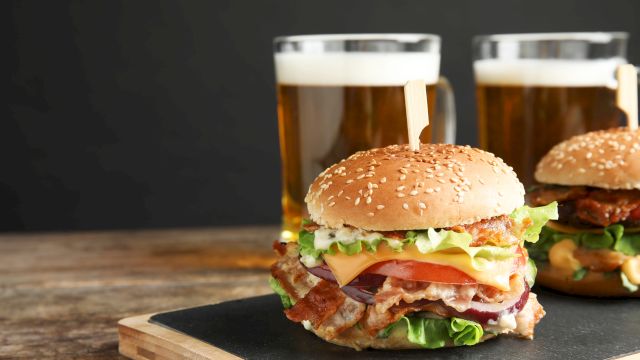 Two burgers with lettuce, tomato, cheese and bacon on a wooden board, plus two mugs of beer in the background.