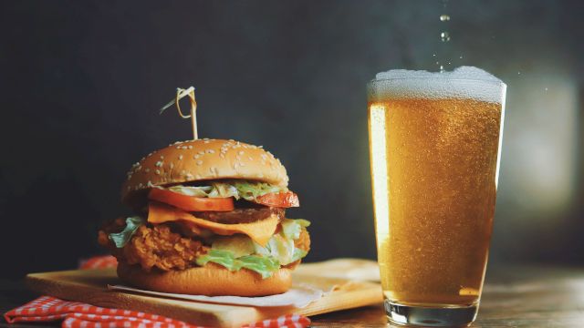 A juicy burger with lettuce, tomato, cheese and pickup fork beside a cold beer on a wooden table.
