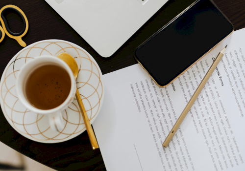 A cup of coffee on a saucer with a spoon, beside a smartphone, scissors, a pencil, and papers on a dark desk.