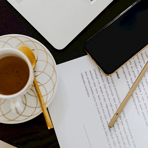 A cup of coffee on a saucer with a spoon, beside a smartphone, scissors, a pencil, and papers on a dark desk.