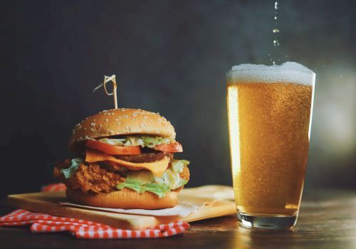 A crispy burger with lettuce, tomato, and cheese on a bun, a wooden board, and a tall glass of beer beside it, all on a dark tabletop.