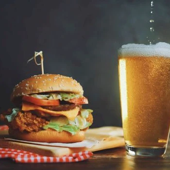 A crispy burger with lettuce, tomato, and cheese on a bun, a wooden board, and a tall glass of beer beside it, all on a dark tabletop.
