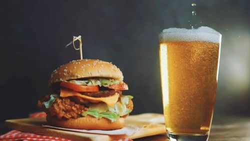 A crispy burger with lettuce, tomato, and cheese on a bun, a wooden board, and a tall glass of beer beside it, all on a dark tabletop.