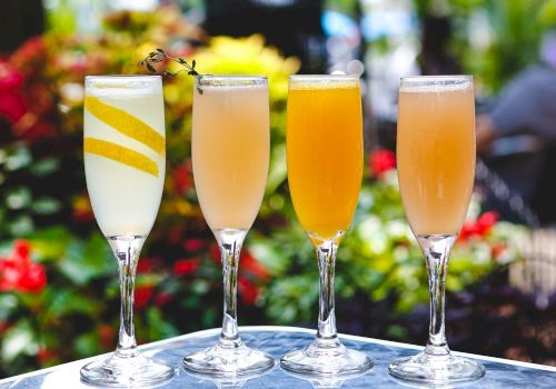 Four champagne flutes with colorful drinks on a sunny outdoor table, garnished and ready to toast.