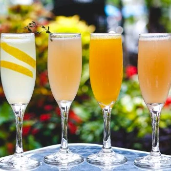 Four champagne flutes with colorful drinks on a sunny outdoor table, garnished and ready to toast.