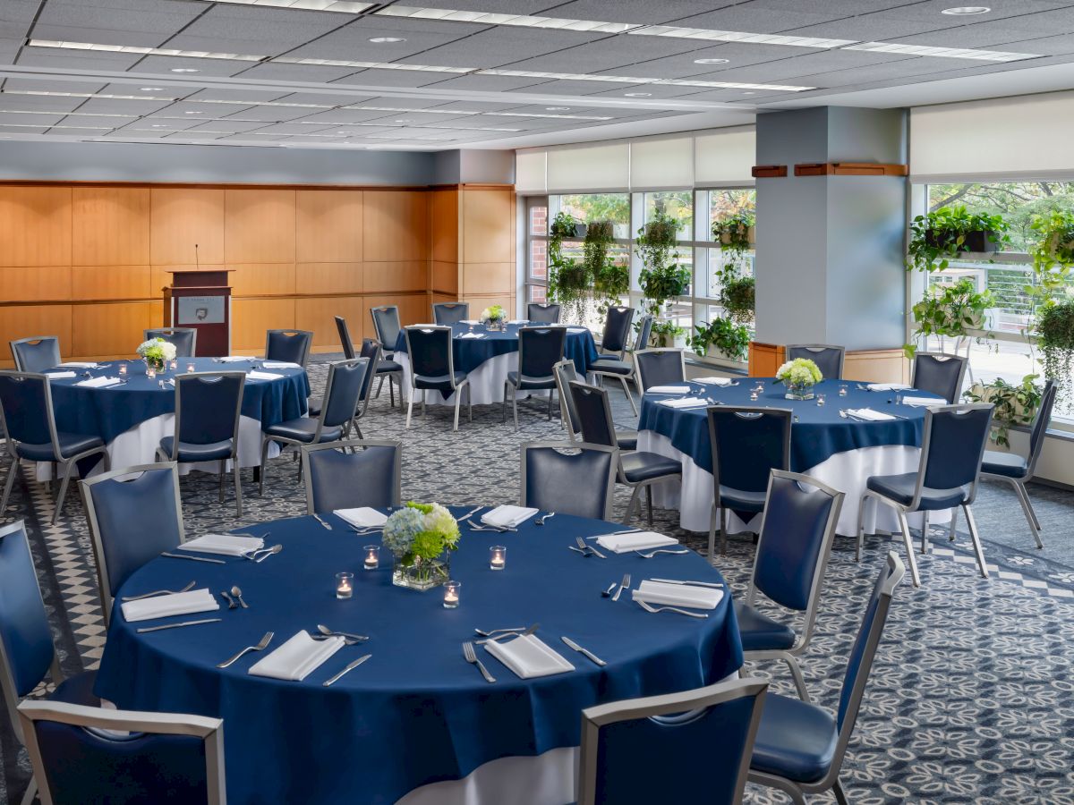A business conference room set with round tables draped in blue cloth, formal place settings, chairs, and potted plants by windows.