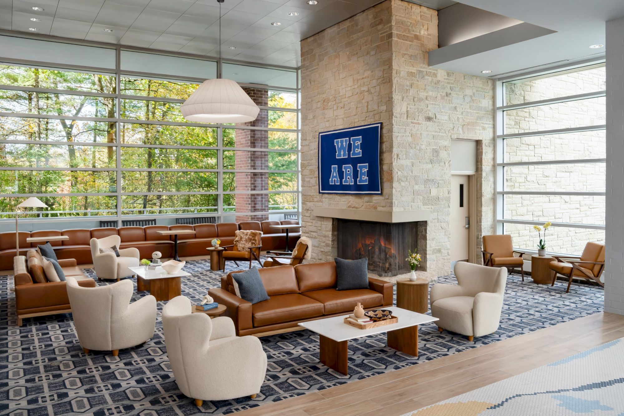 A spacious hotel lobby with tan leather sofas, cream chairs, wooden coffee tables, a stone fireplace, large windows, and a blue geometric rug&mdash;bright and modern.